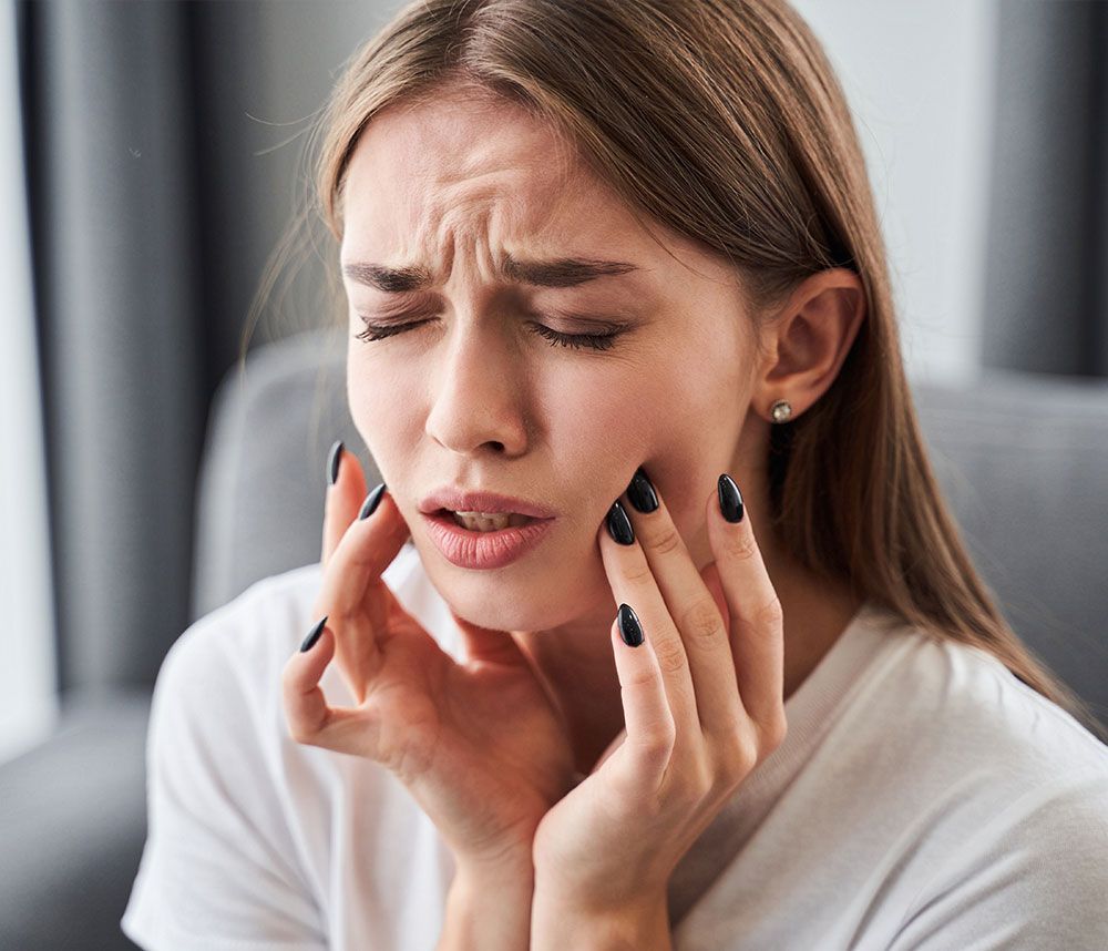 Frau mit schmerzverzerrtem Gesicht hält sich beide Wangen wegen Zahnschmerzen.